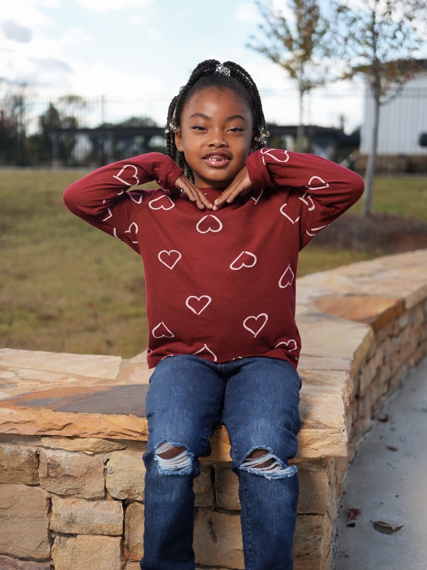 A young girl in a burgundy heart-print top smiling while seated on a stone wall