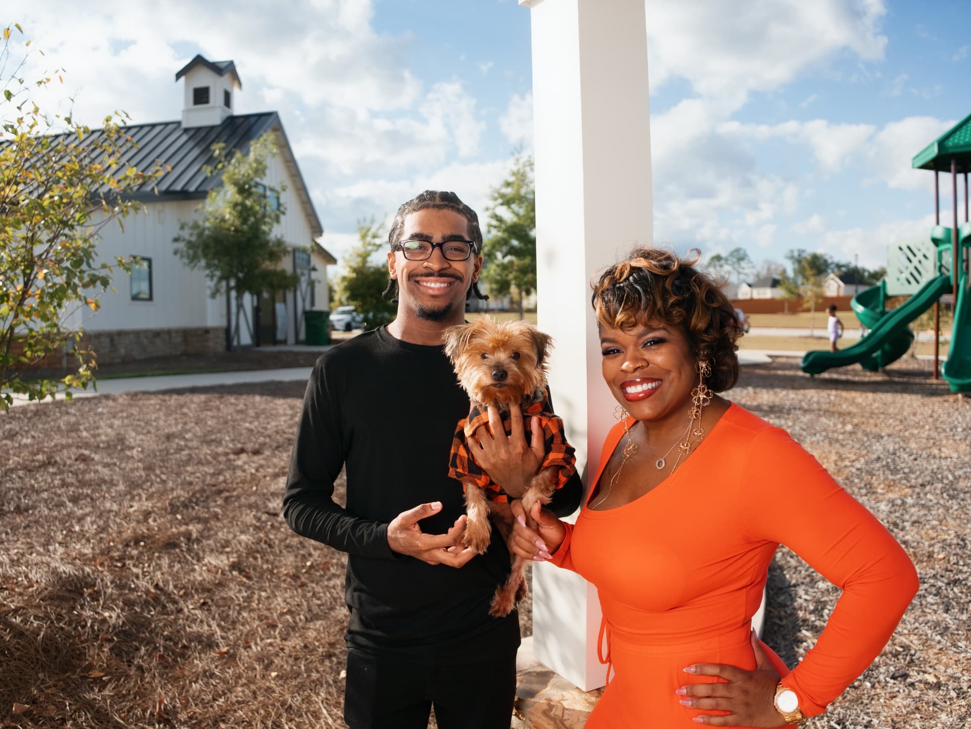 A mother and her son holding their small dog beside a community clubhouse and playground