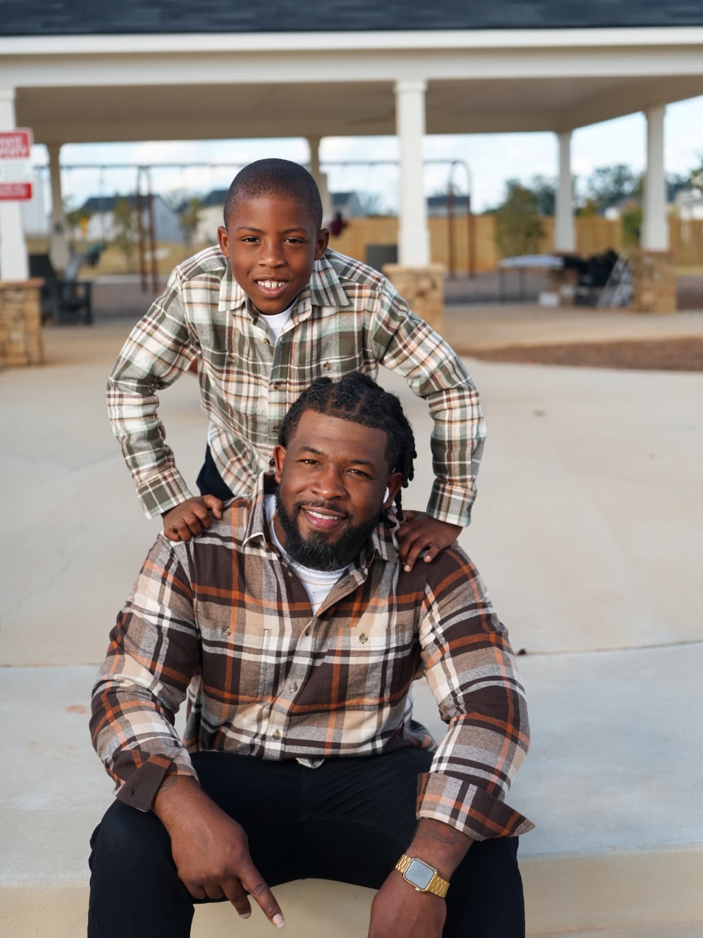 A father seated outdoors with his son leaning over his shoulders, both wearing flannel shirts